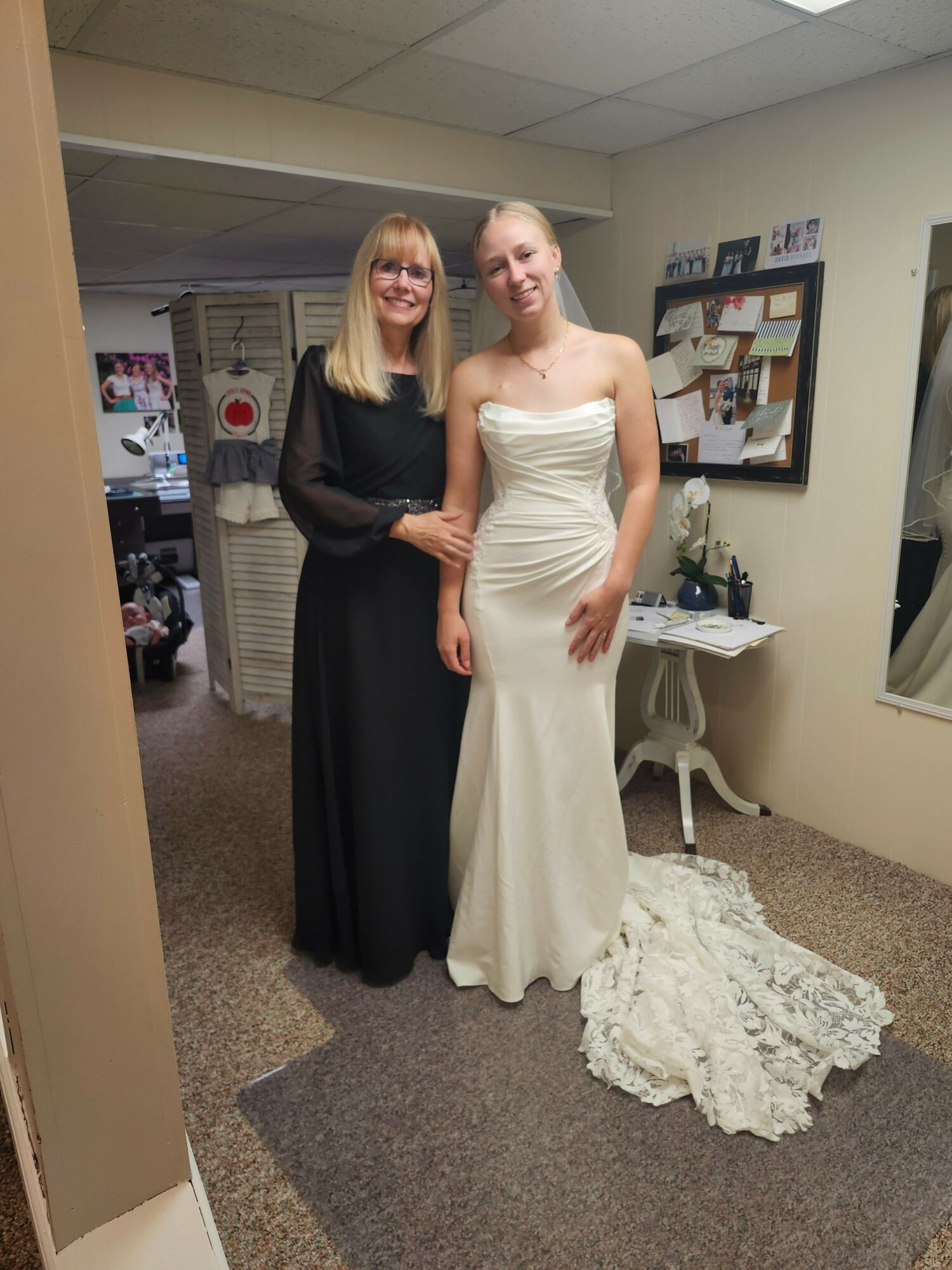 Two women, one in a white wedding dress and the other in a black dress, pose together indoors. The woman in white is smiling, and the woman in black is holding her arm. A white dress is on the floor nearby.