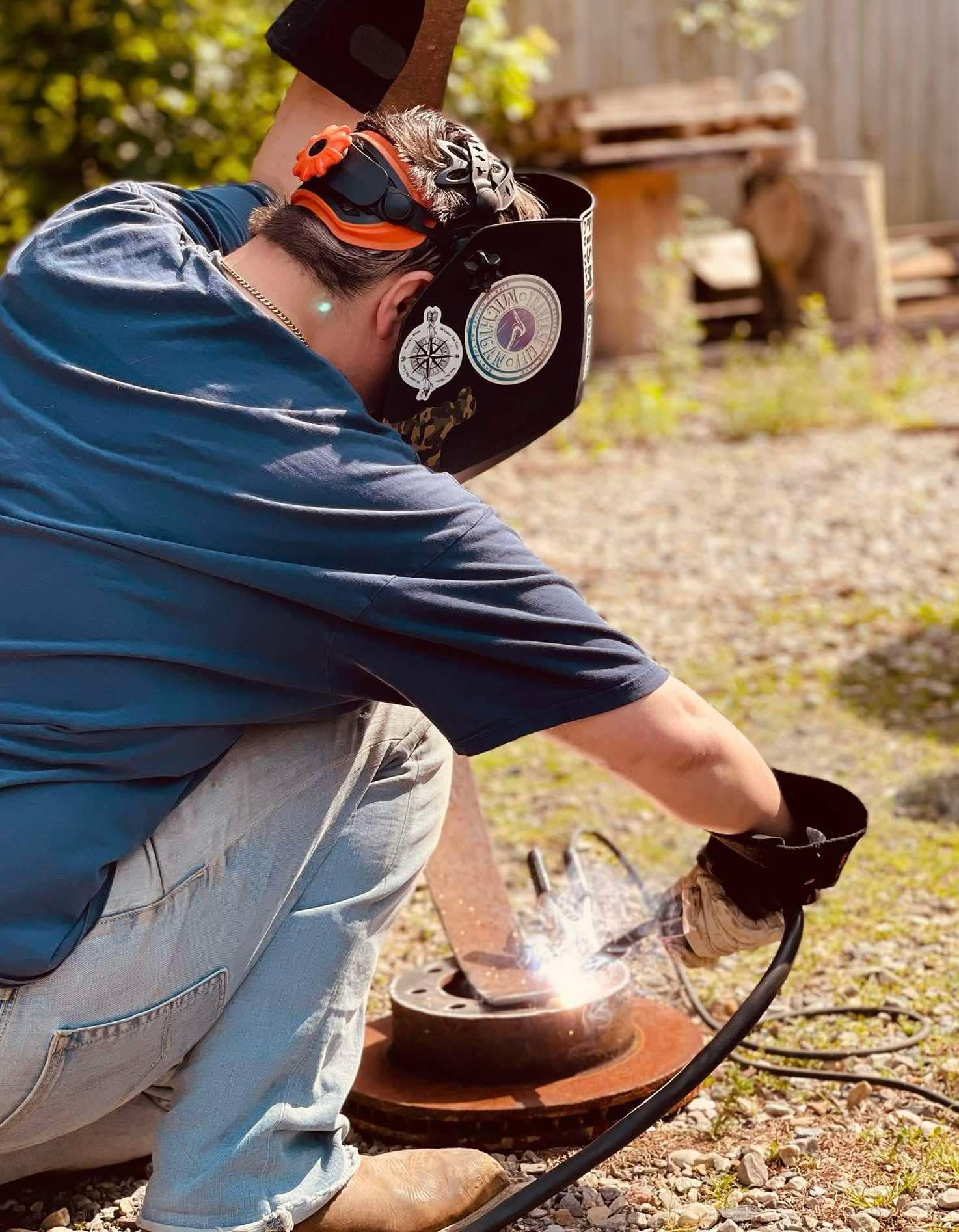 Person wearing protective gear welds metal outdoors, sparks flying from the welding process, with a wooden fence and trees in background.