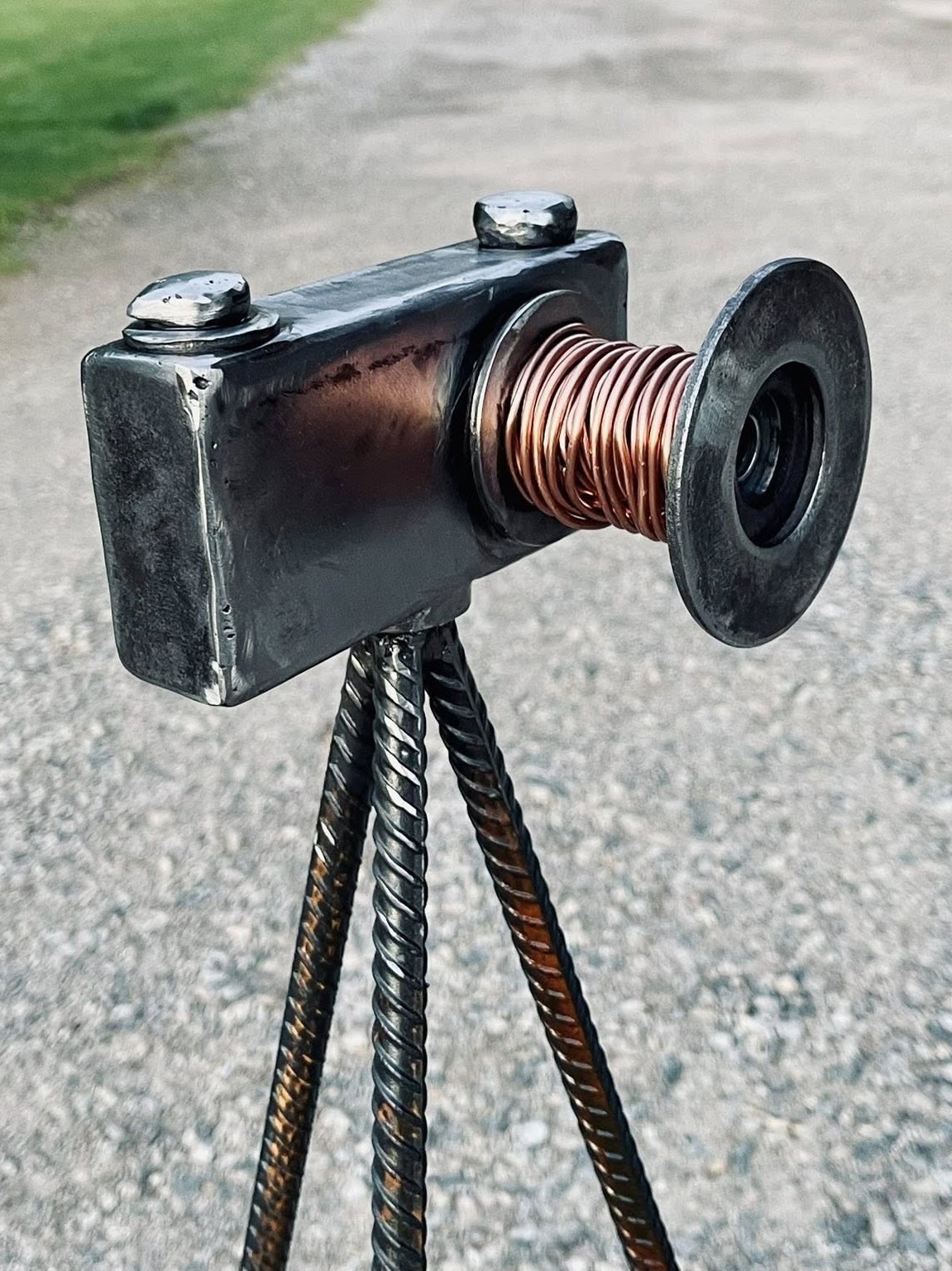 Object resembling a camera or device with a black rectangular body, a large spool, and three legs, outdoors on a gravel surface.