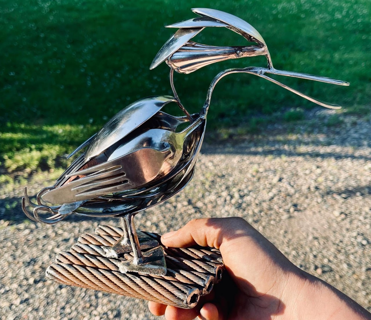 Hand holding a shiny metal bird sculpture with a long beak and detailed feathers, outdoors on a gravel surface.