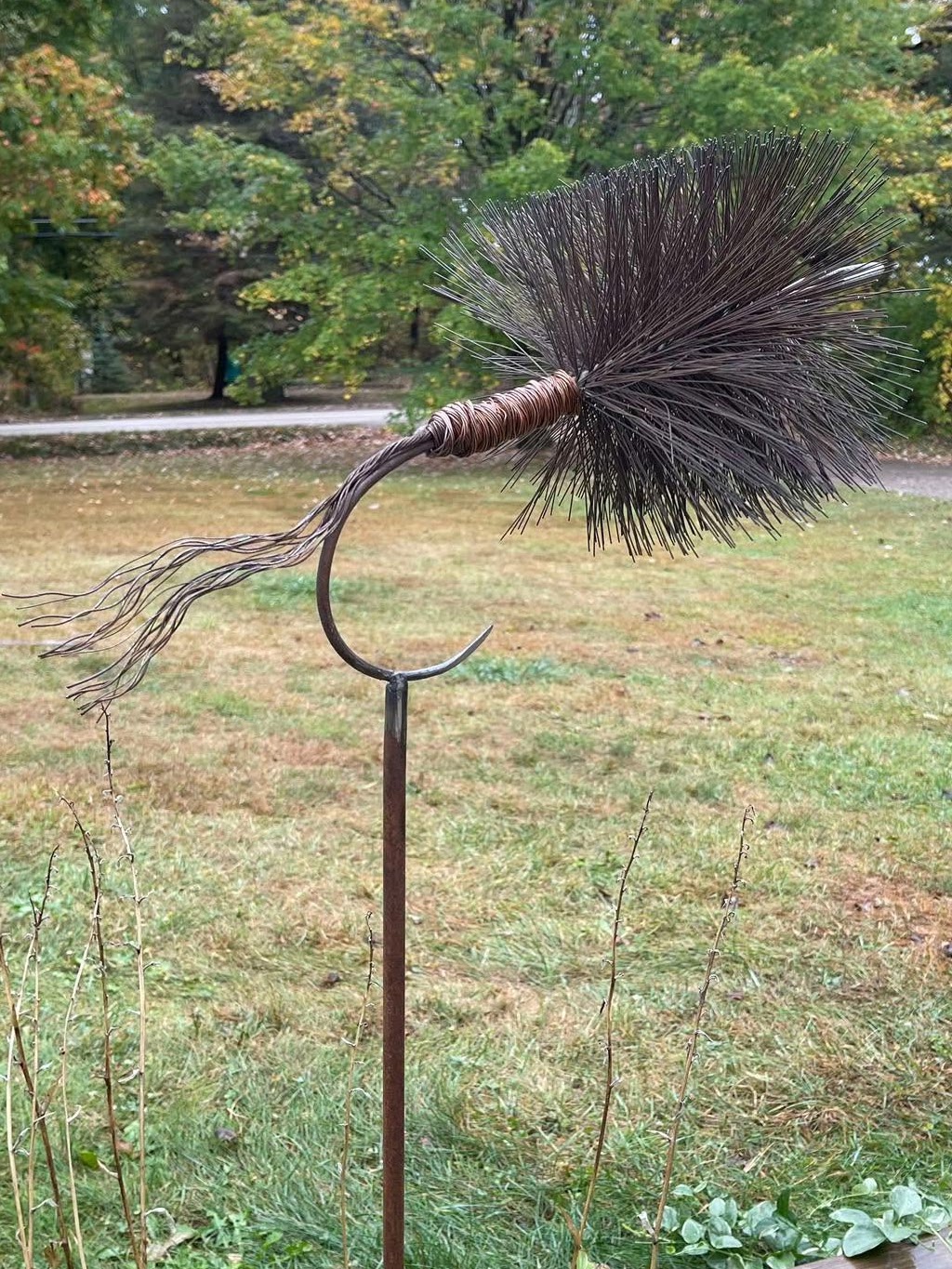 A fishing lure with a black feathered tail and a curved metal hook, mounted on a stand outdoors with trees and grass.