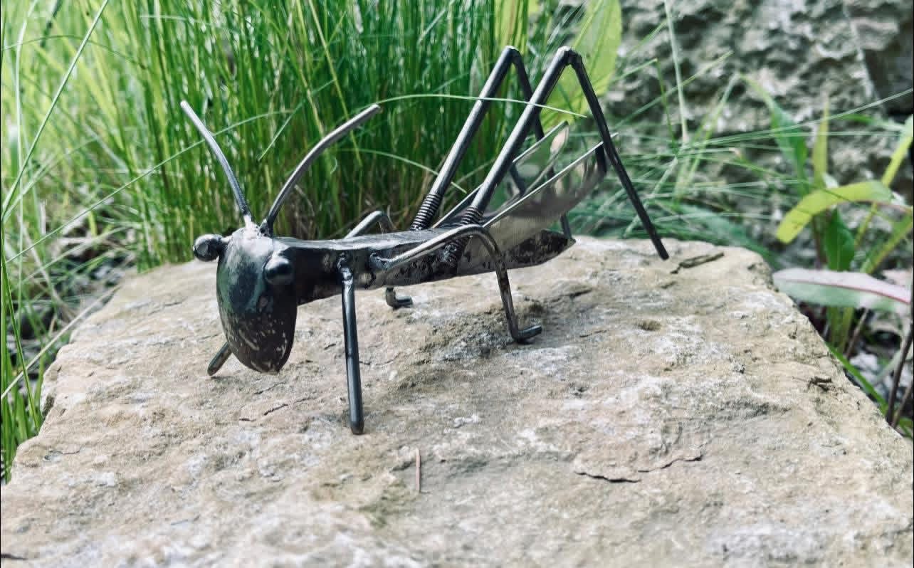 A realistic-looking insect sculpture on a rock with grass and plants in the background.