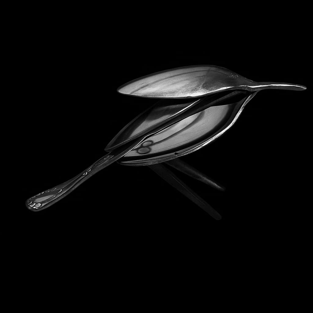 A metallic spoon with a leaf-shaped bowl, placed on a reflective black surface, with a black background.