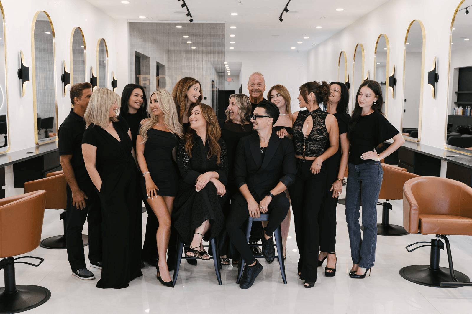Group of people in a salon, some sitting and some standing, with mirrors and chairs in the background.