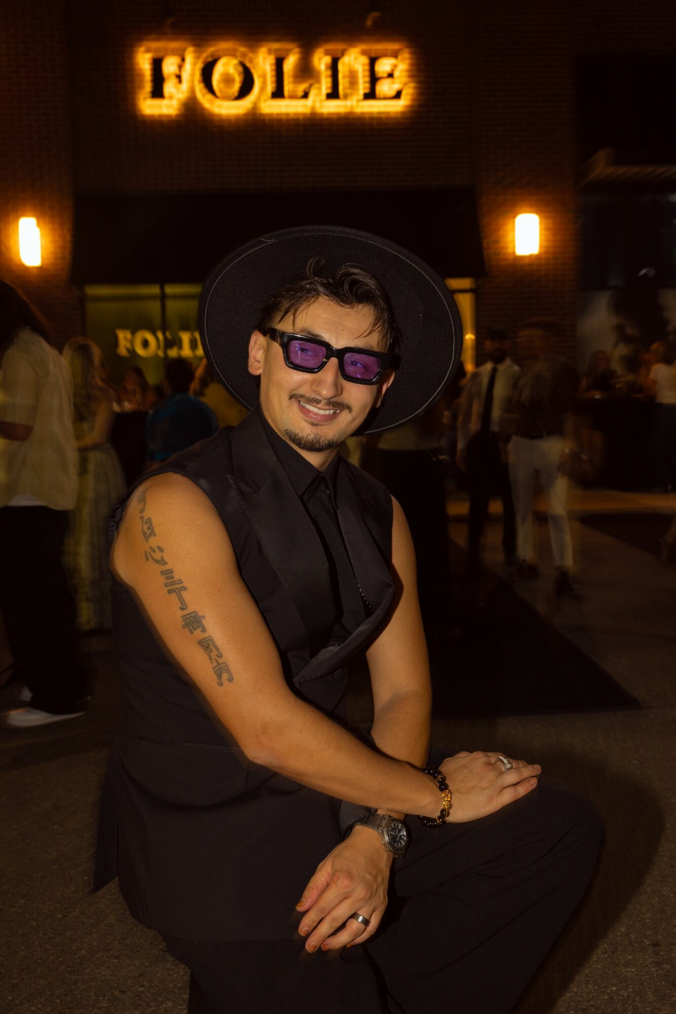 Person wearing sunglasses and a hat, smiling, sitting with crossed legs in front of a lit sign that says 'HOLLE'.