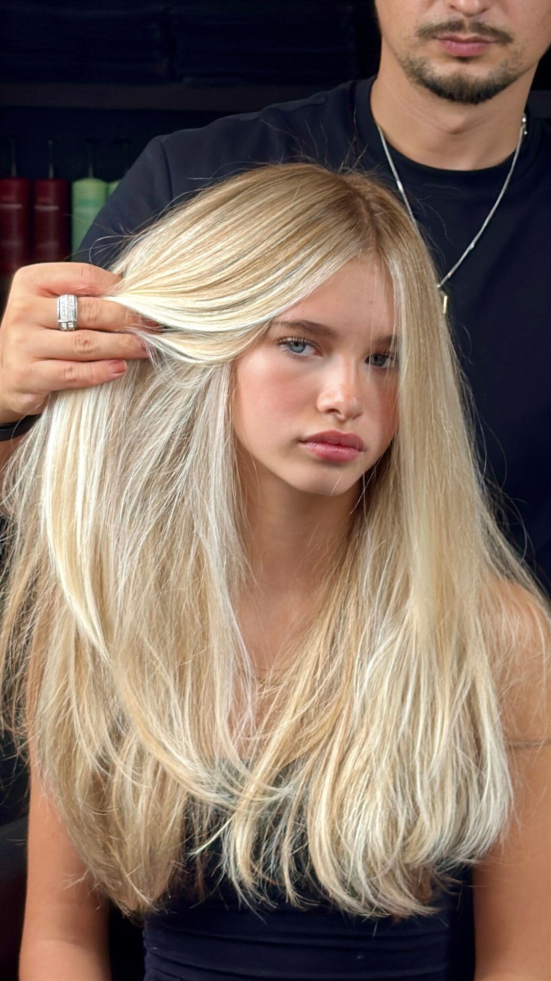 Hairdresser styles a young woman's long blonde hair in a salon setting.