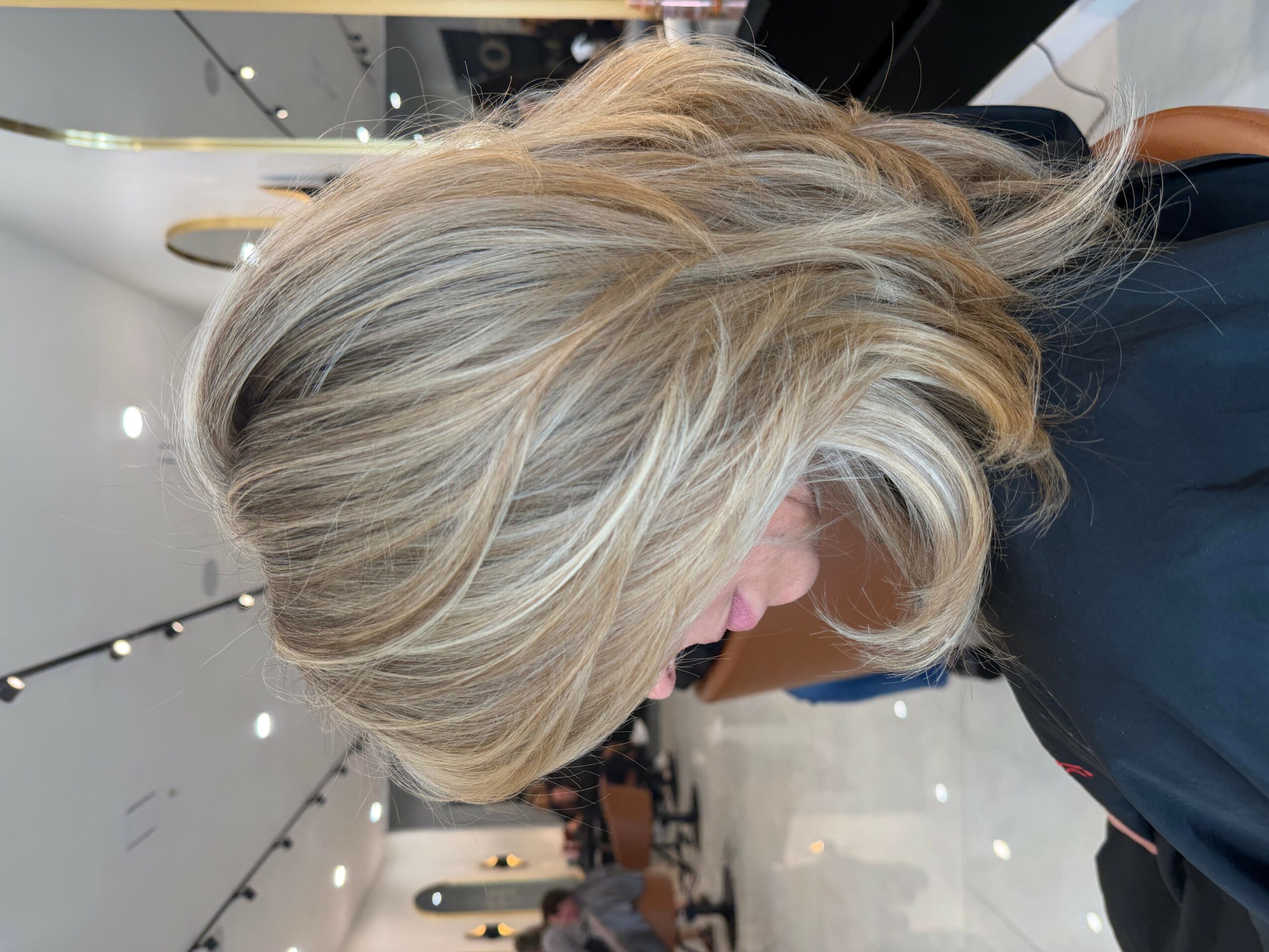 Side view of a woman with short, layered blonde hair in a salon, with salon equipment in the background.