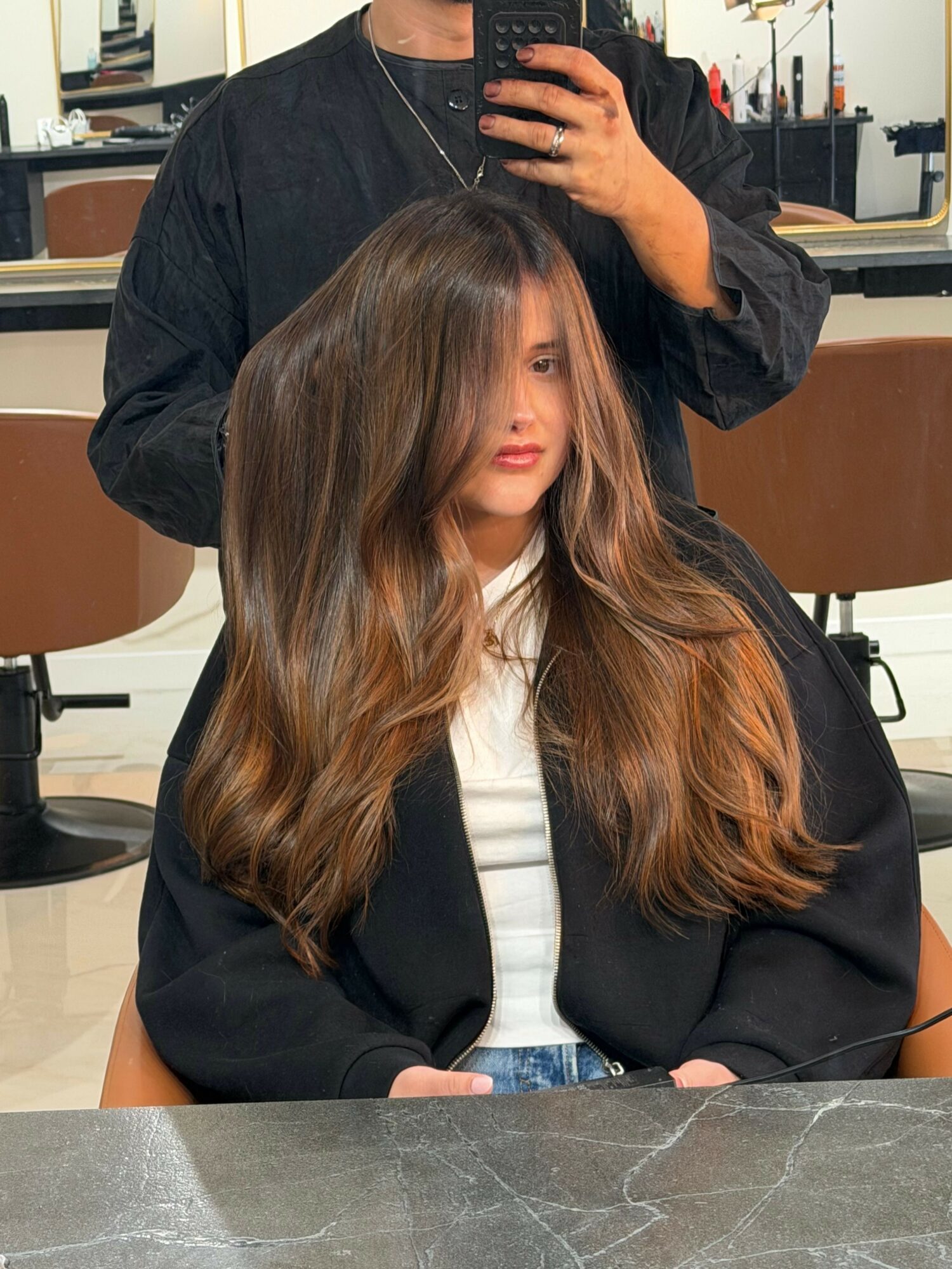 Young woman with long, wavy hair sitting at a table, looking at the camera, in a casual setting.