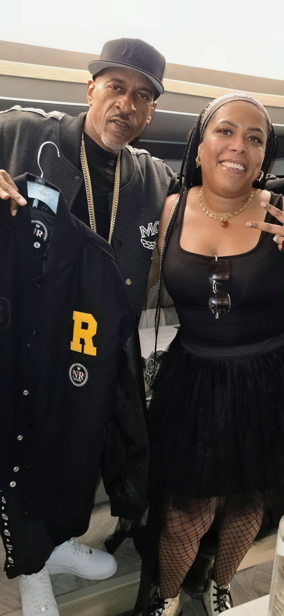 Two people standing indoors, one holding a black jacket with a yellow letter R, smiling, with a woman wearing a black dress and fishnet stockings.