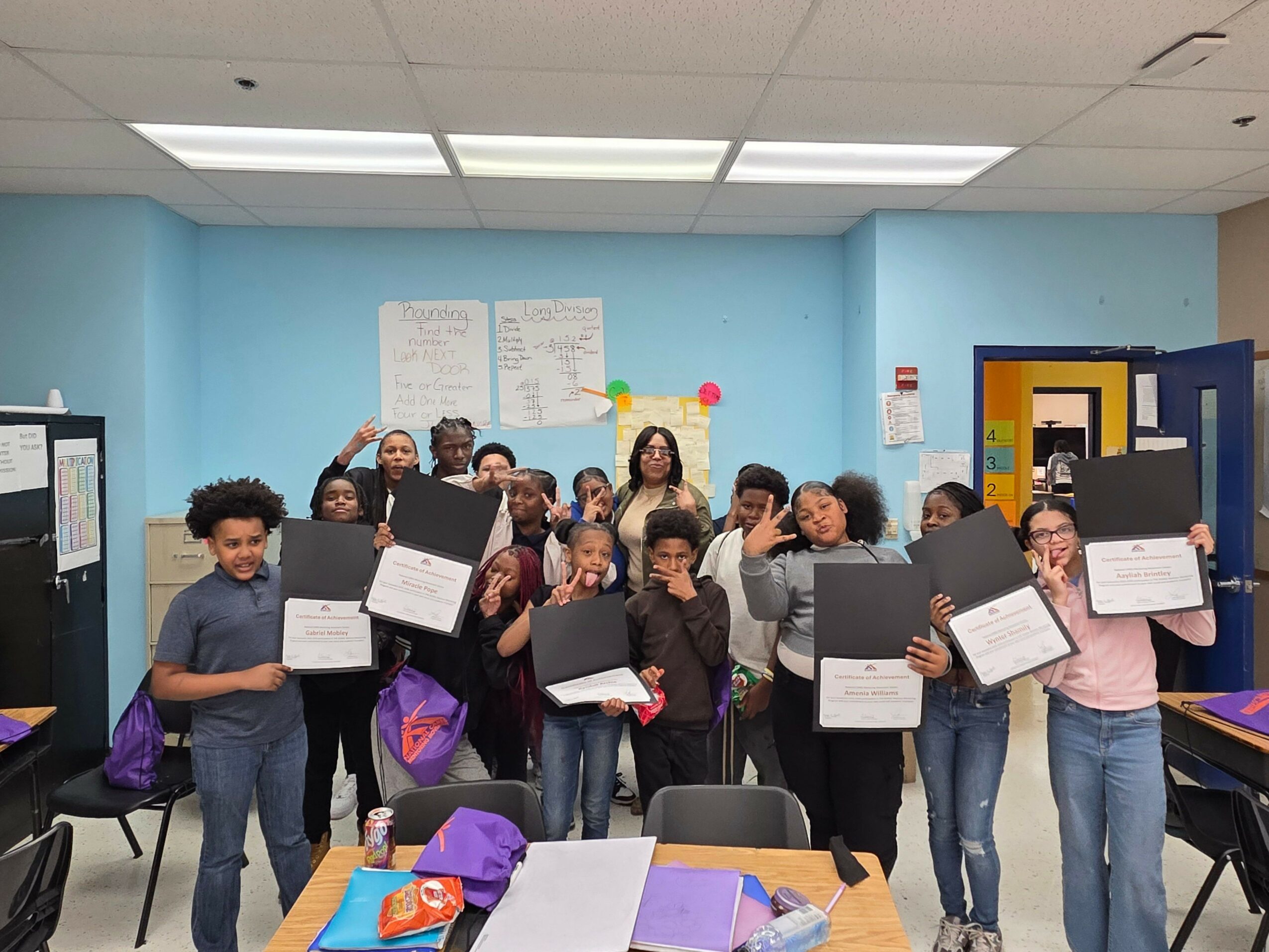 Group of people holding certificates and gift bags in a classroom with blue walls and windows.