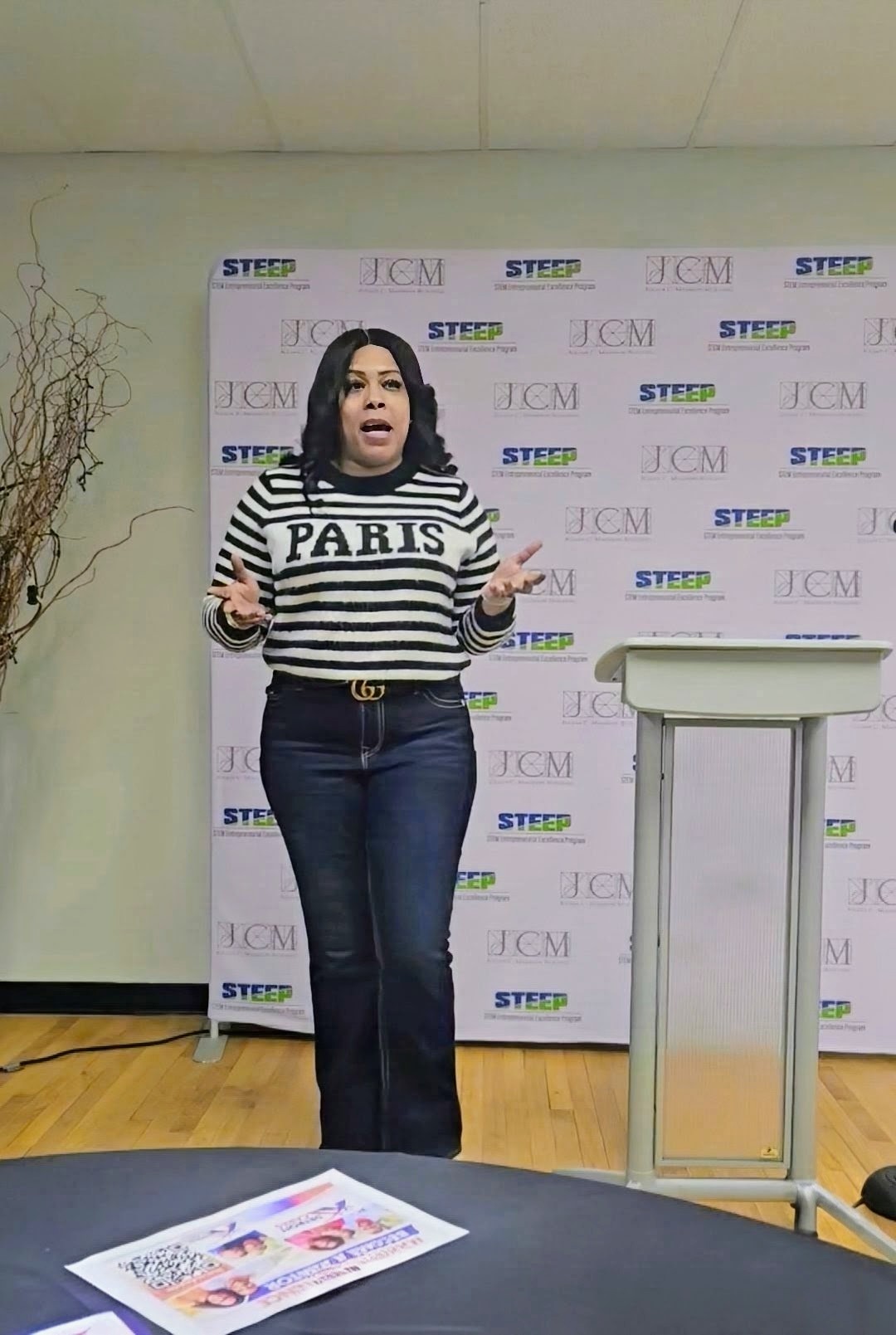 Woman with shoulder-length dark hair speaking in front of a backdrop with logos, wearing a striped sweater and jeans.