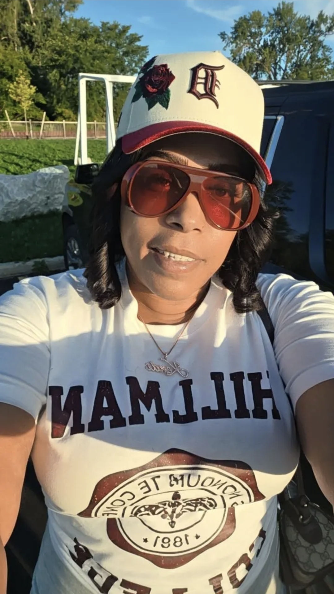 Woman wearing a white baseball cap, sunglasses, and a white T-shirt outdoors with trees in background.