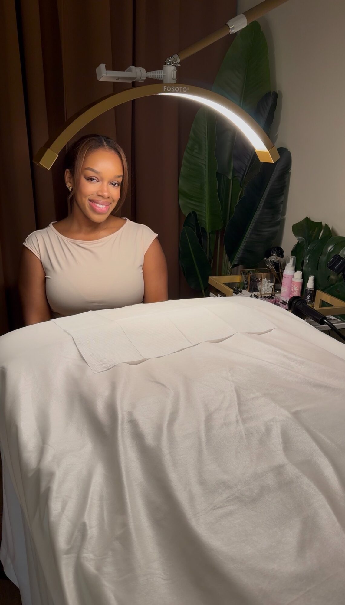 Smiling woman sitting in a room with a bed, plants, and a lamp overhead.
