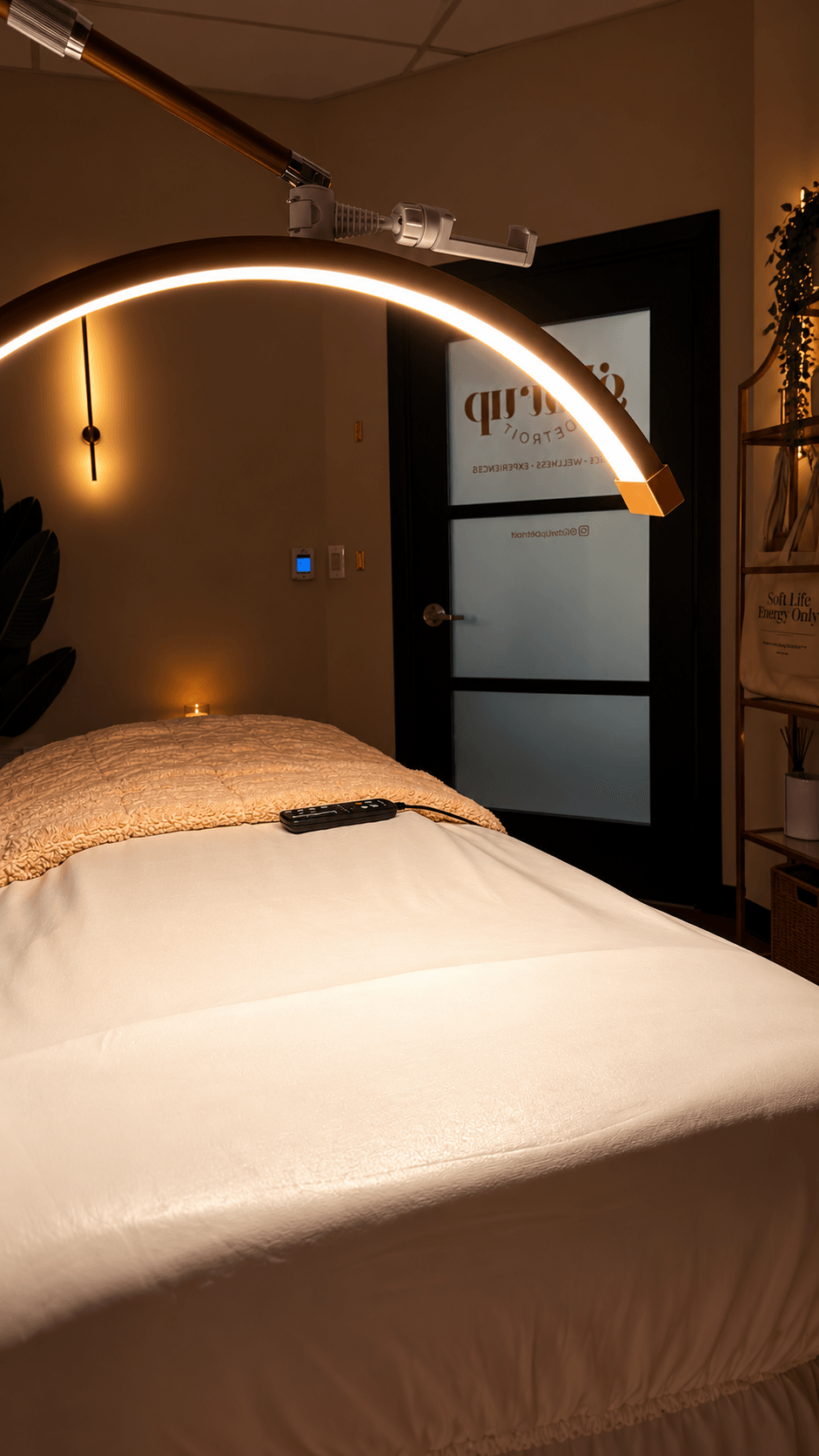A bed with a beige pillow and a remote control on white sheets, illuminated by a curved LED light fixture, in a room with a door and wall decor.