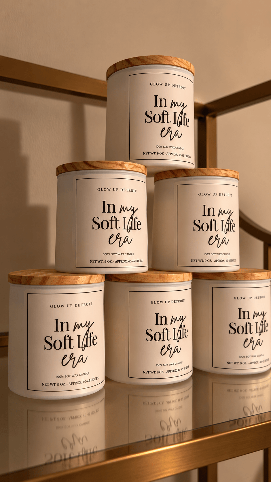 Six white containers with wooden lids stacked on a glass shelf, labeled 'In my Soft Life era,' in a room with beige walls.