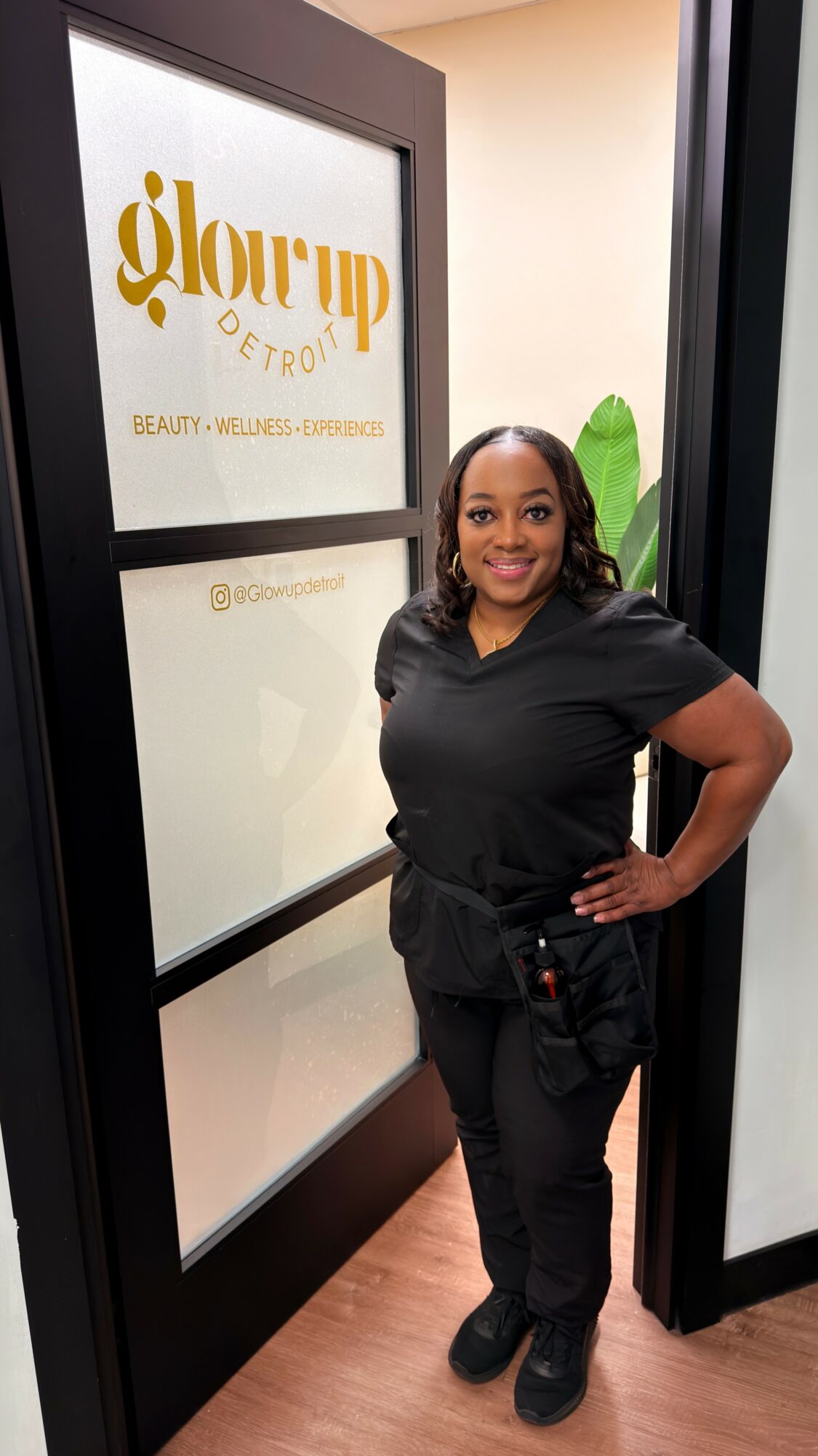 Smiling woman in black scrubs standing inside a beauty salon, with a sign on the door reading 'glow up' and 'beauty'.