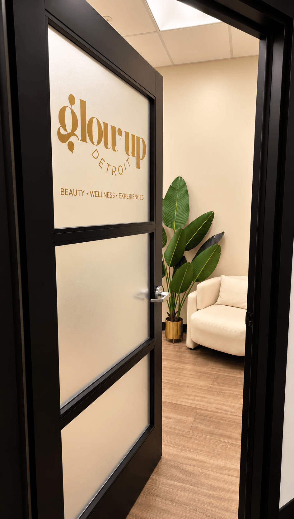 View through a glass door into a room with a white sofa, large green plant, and a sign reading 'glow up' on the wall.