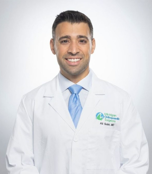 Man in white coat with blue tie smiling, standing against a plain background.