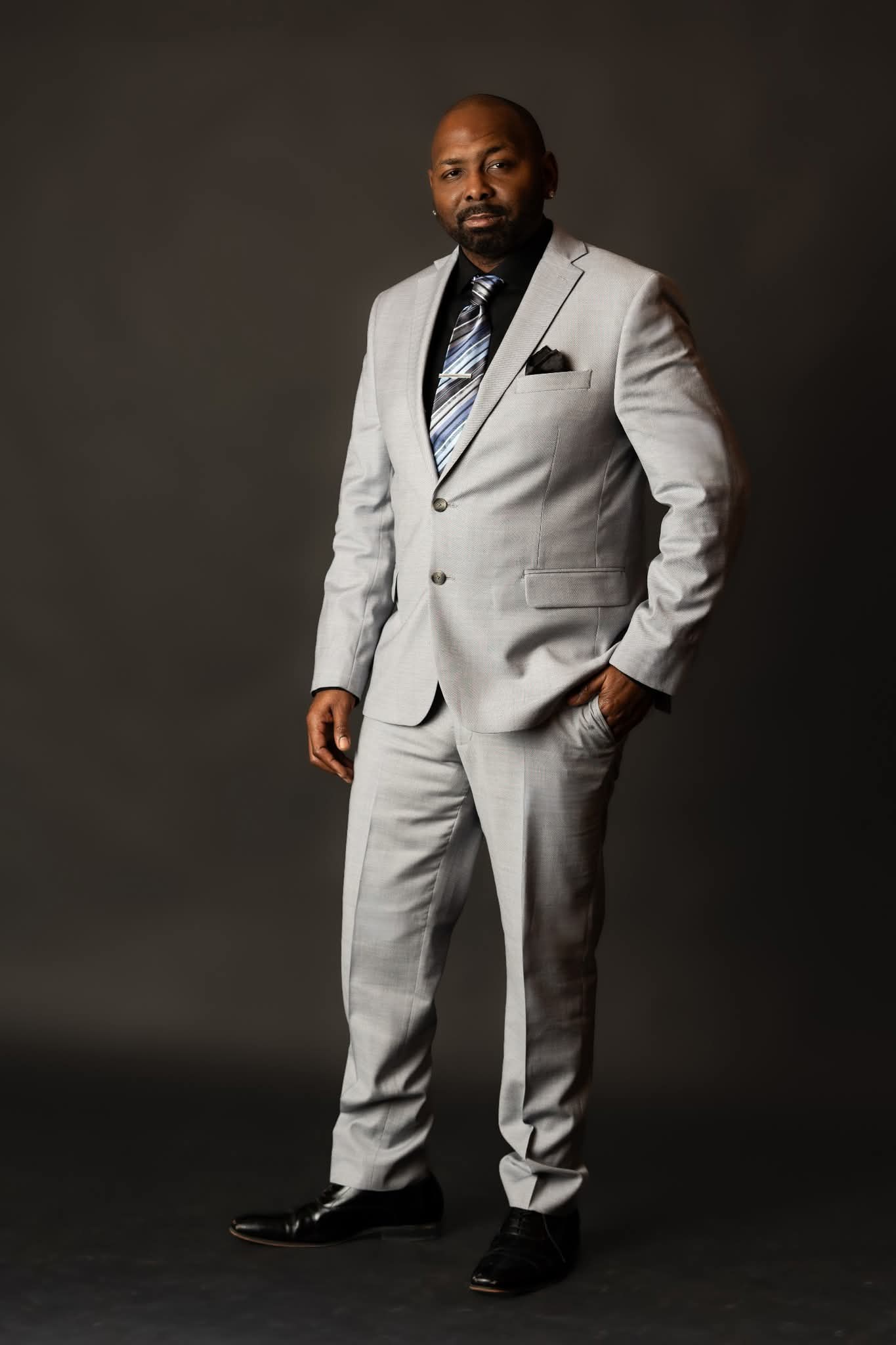 Man in light gray suit with black shirt and patterned tie, standing with hand in pocket, against dark background.