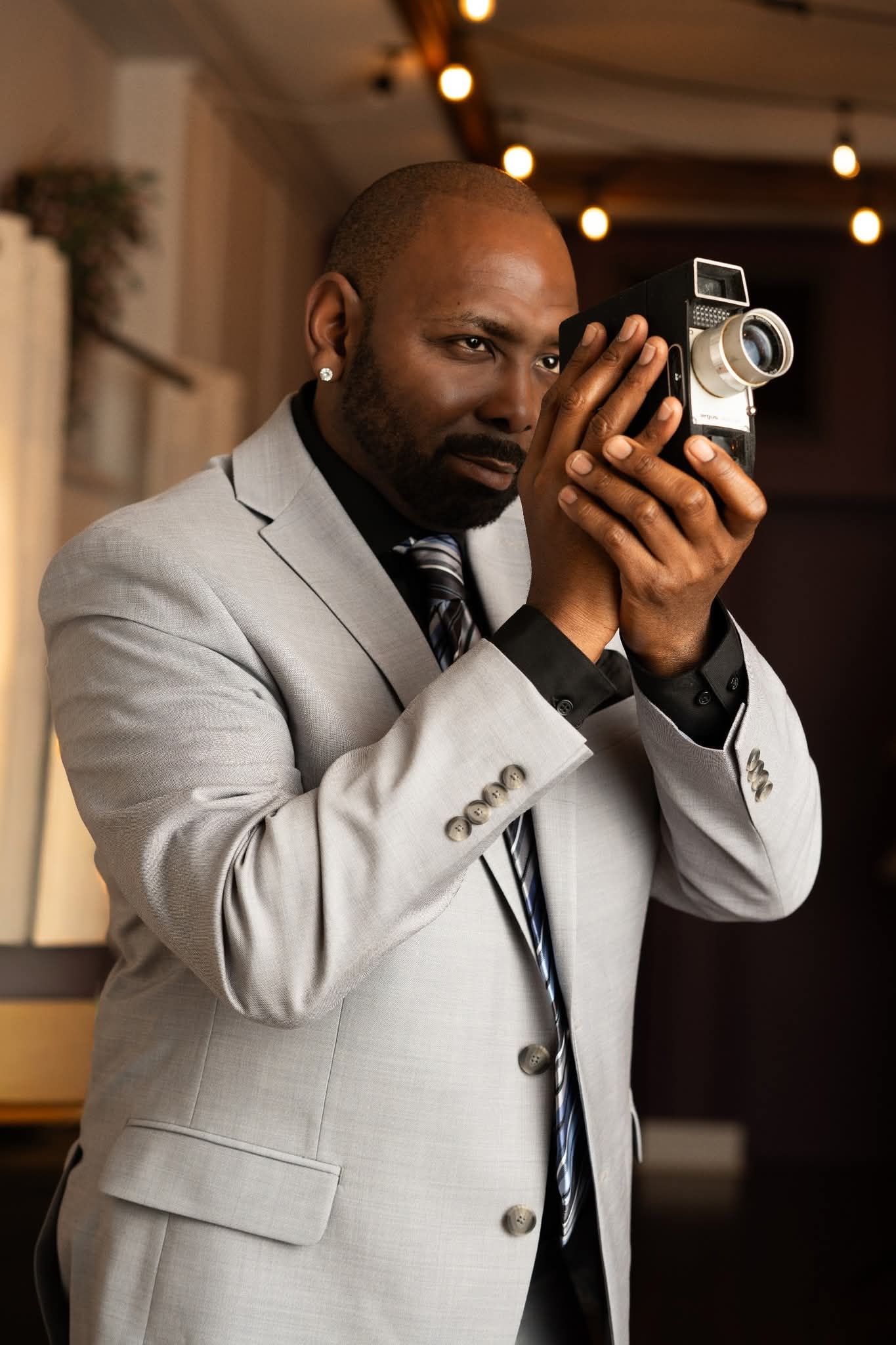 Man in a light gray suit holding a vintage camera indoors, with warm lighting and blurred background.
