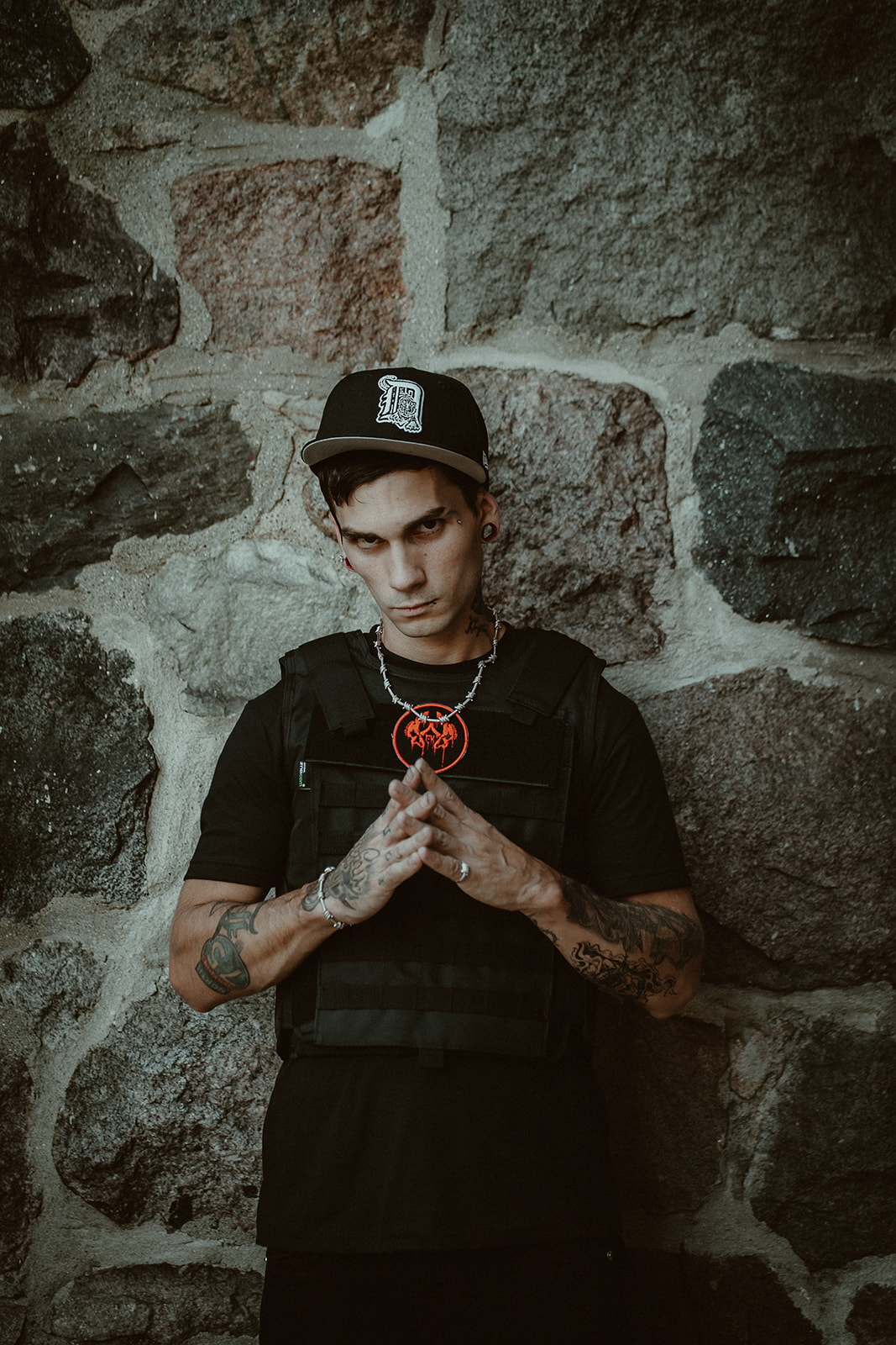 Young person with tattoos and a black cap stands against a stone wall, holding a necklace with both hands.