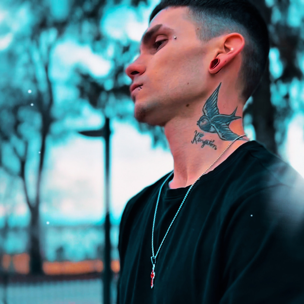 Side profile of a person with a tattoo of a bird on the neck, wearing a black shirt and a necklace, outdoors.