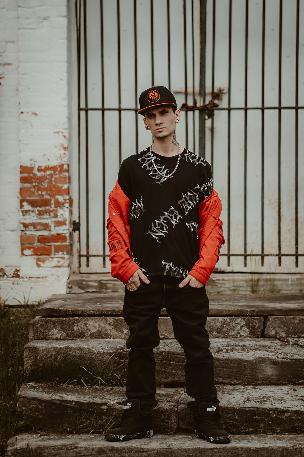 Young man standing outdoors in front of a metal gate, wearing a black and red jacket, black pants, and a cap.