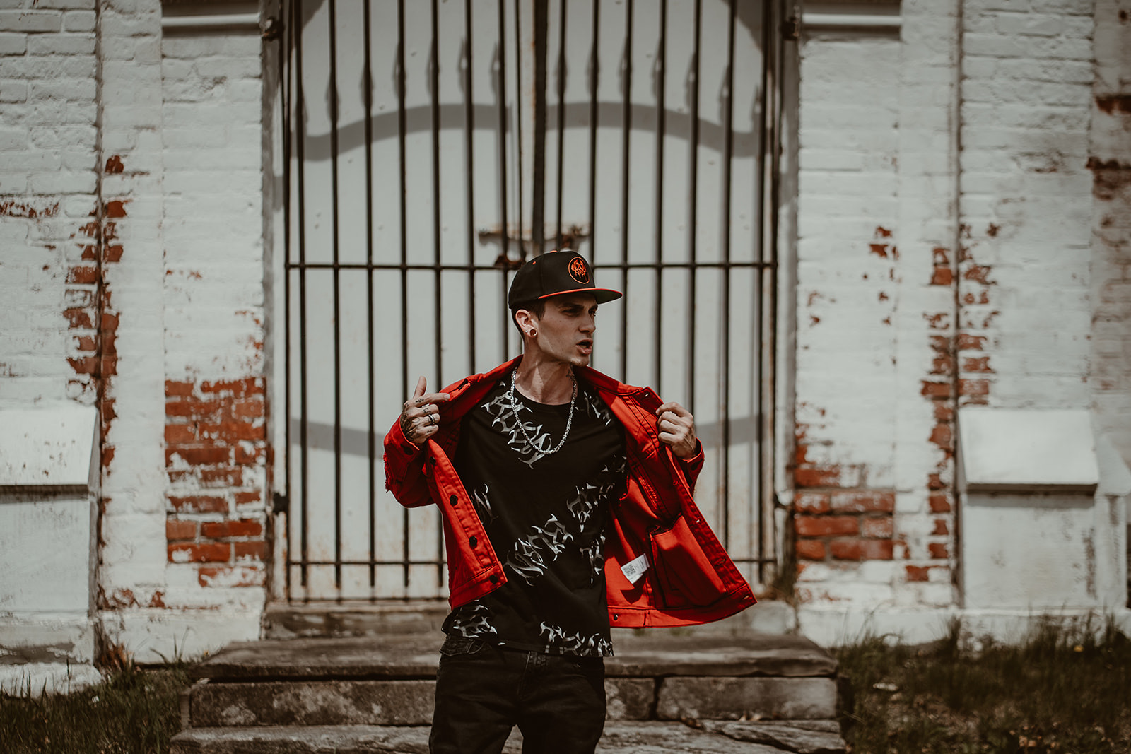 Person wearing a black shirt and red jacket standing in front of a metal gate and brick wall.