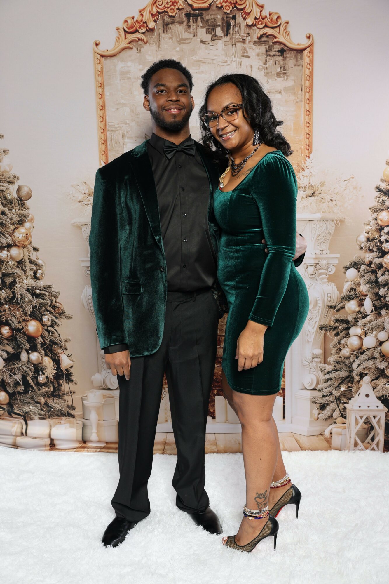 Two women stand indoors in front of a decorated wall, one in a dark outfit and the other in a green velvet dress, both smiling.