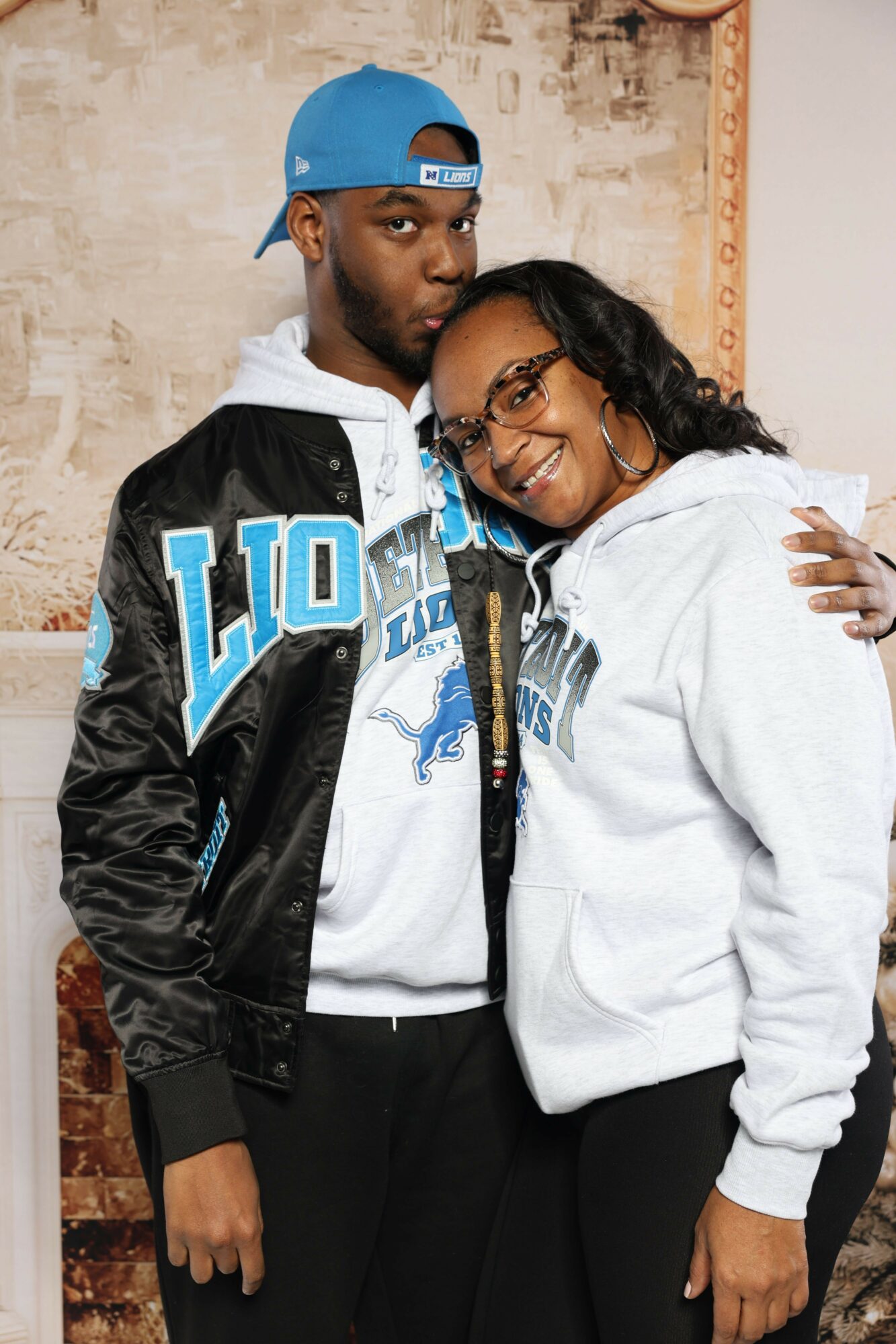 Two people, one wearing a blue cap and black jacket, the other in a white hoodie, pose together indoors.