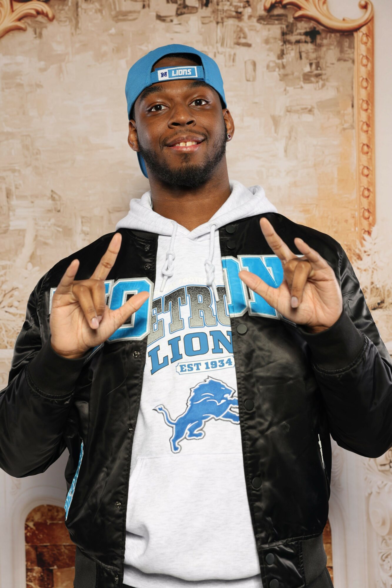 Man wearing a blue cap and black jacket making peace signs with both hands, smiling, in front of a beige wall.