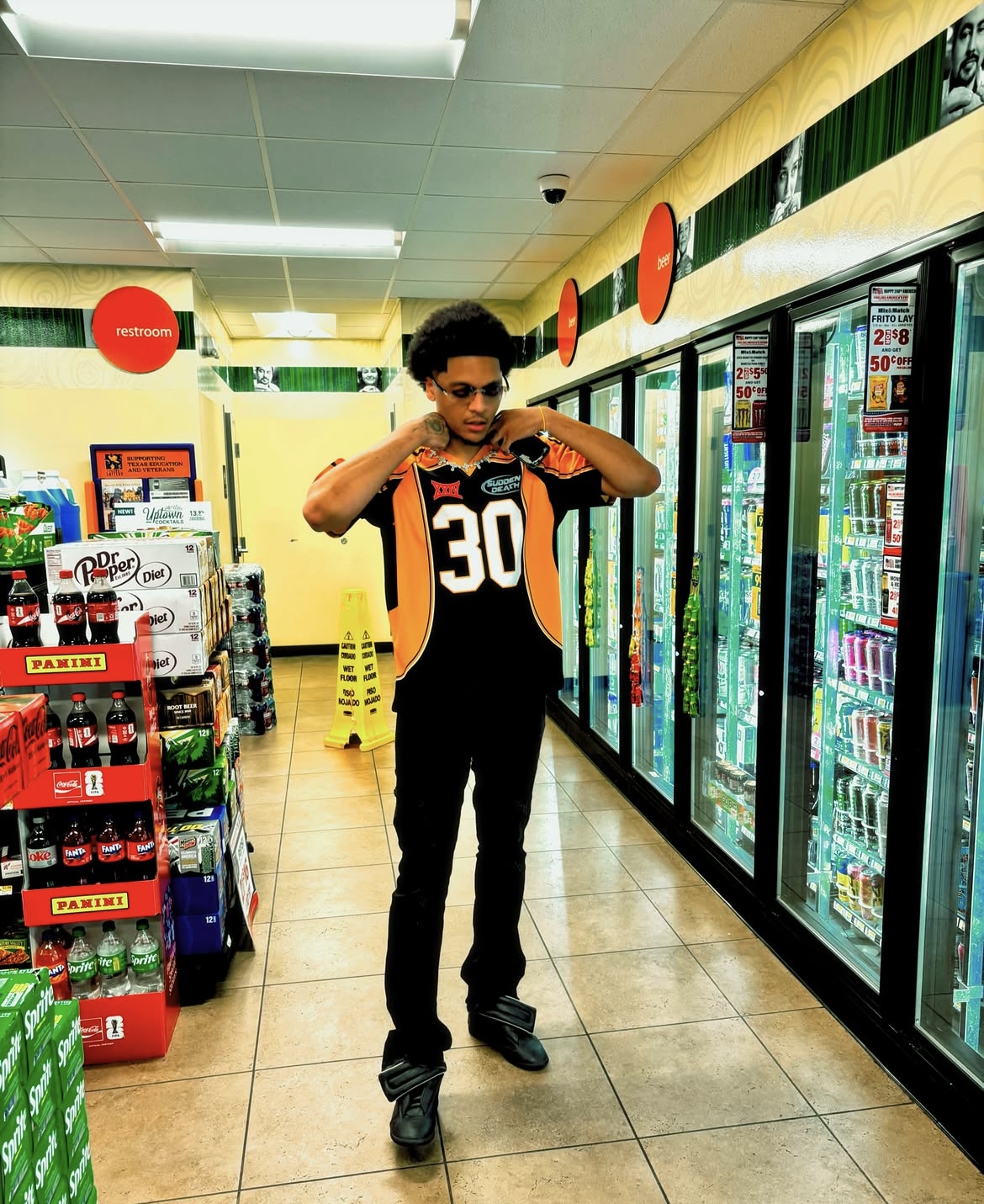 Young person adjusting their black and orange sports jersey inside a grocery store aisle with refrigerated drinks, shelves, and signs.