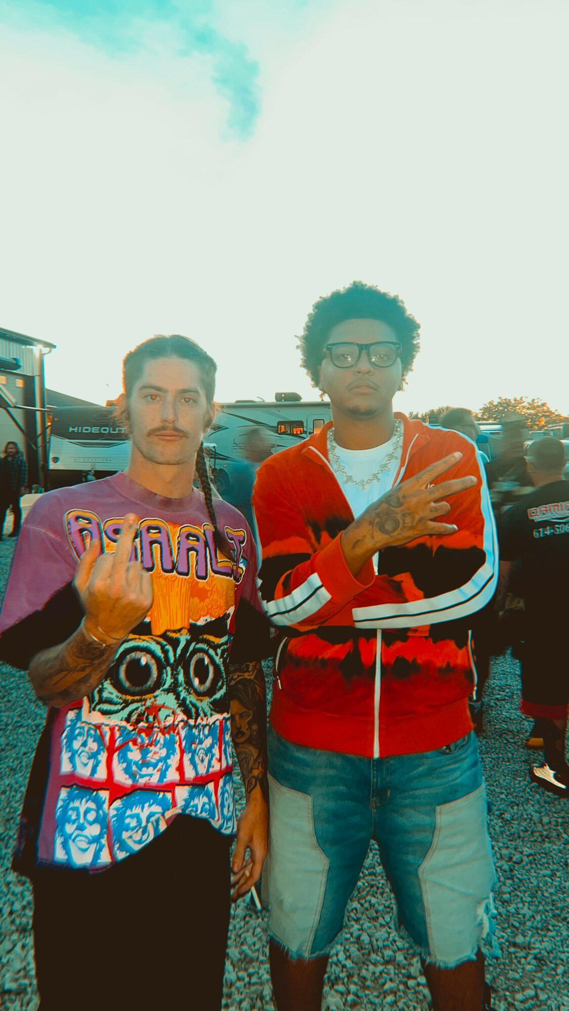 Two young men standing outdoors, one wearing a colorful shirt and the other in a red jacket, with cars in the background.