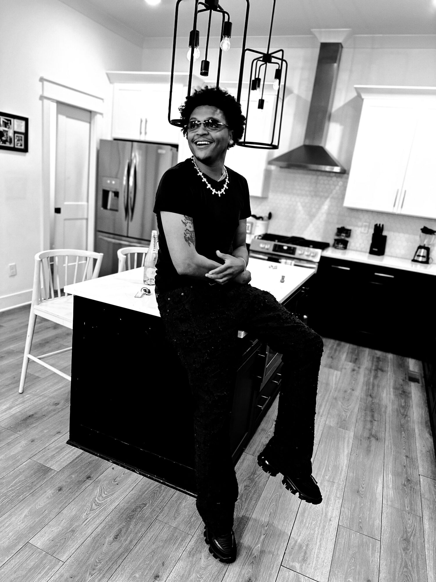 Woman sitting on kitchen island, smiling, in a modern kitchen with pendant lights, white cabinets, and wooden floor.