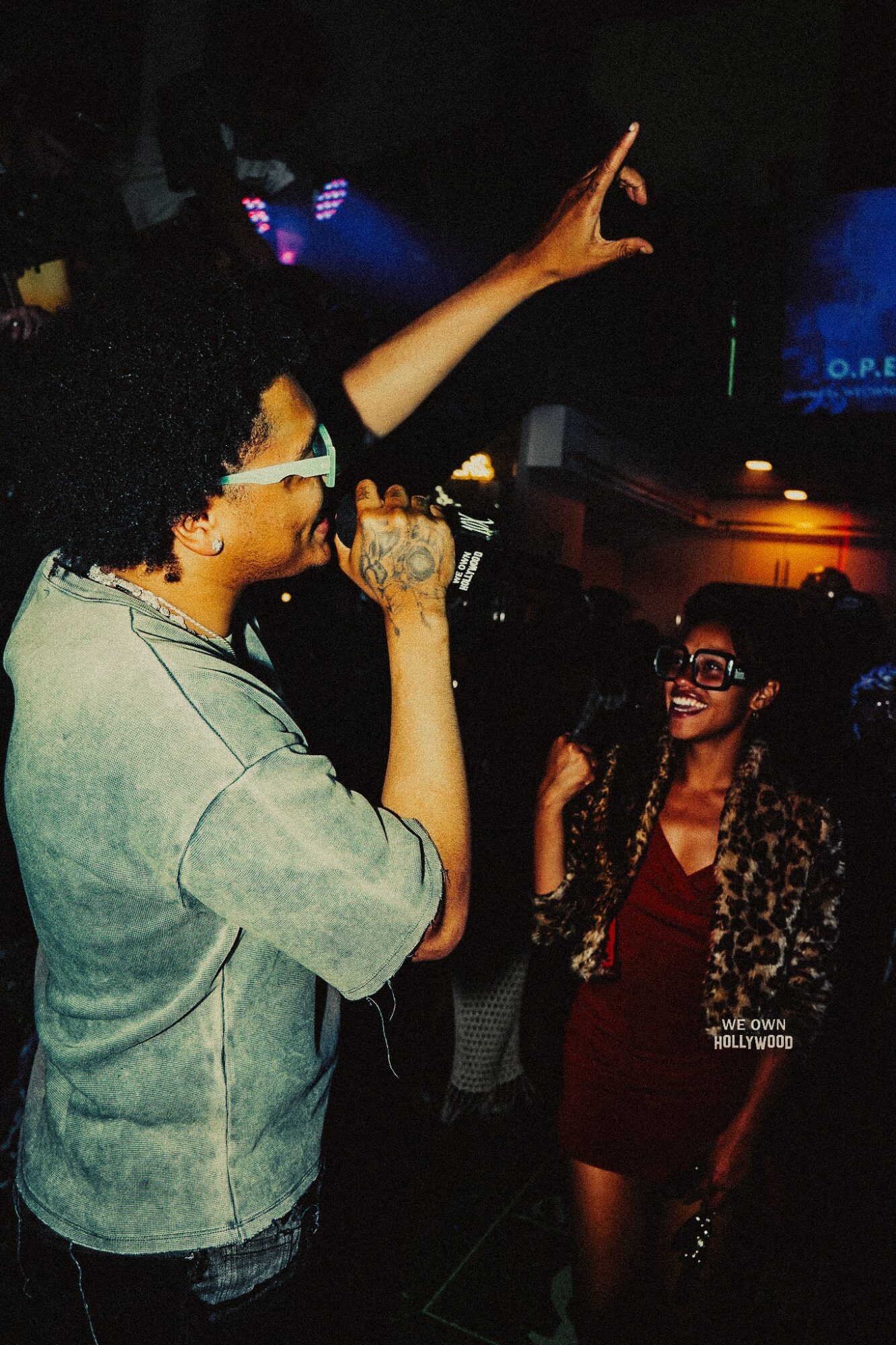 Person with curly hair and glasses singing into a microphone, raising one arm, at a lively event with a smiling woman nearby.