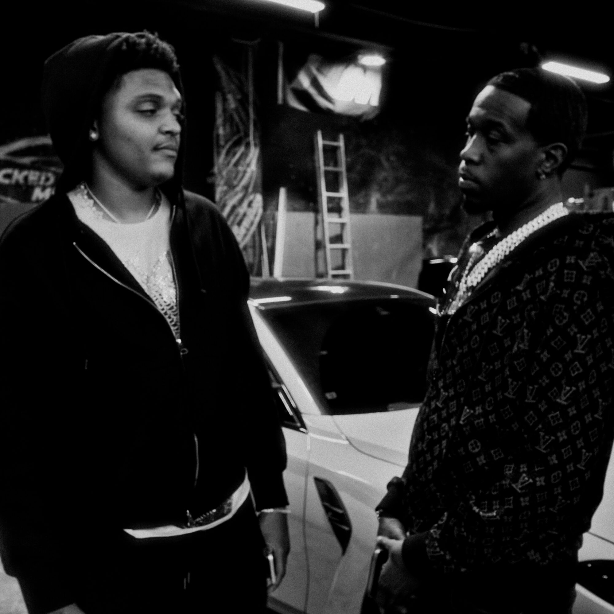 Two young men face each other in a parking lot, with cars and a ladder in the background.