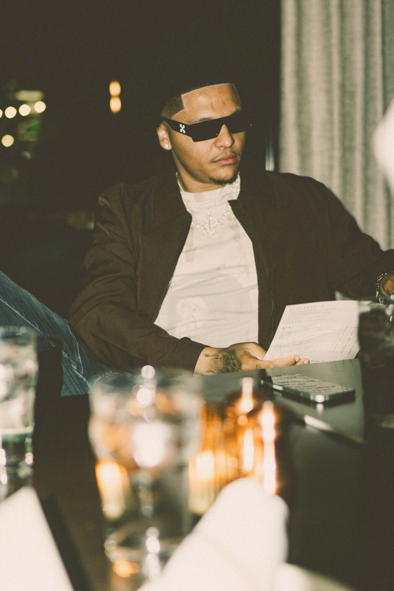 Person wearing sunglasses and a dark jacket, sitting at a table with drinks, holding a paper, in a dimly lit room.