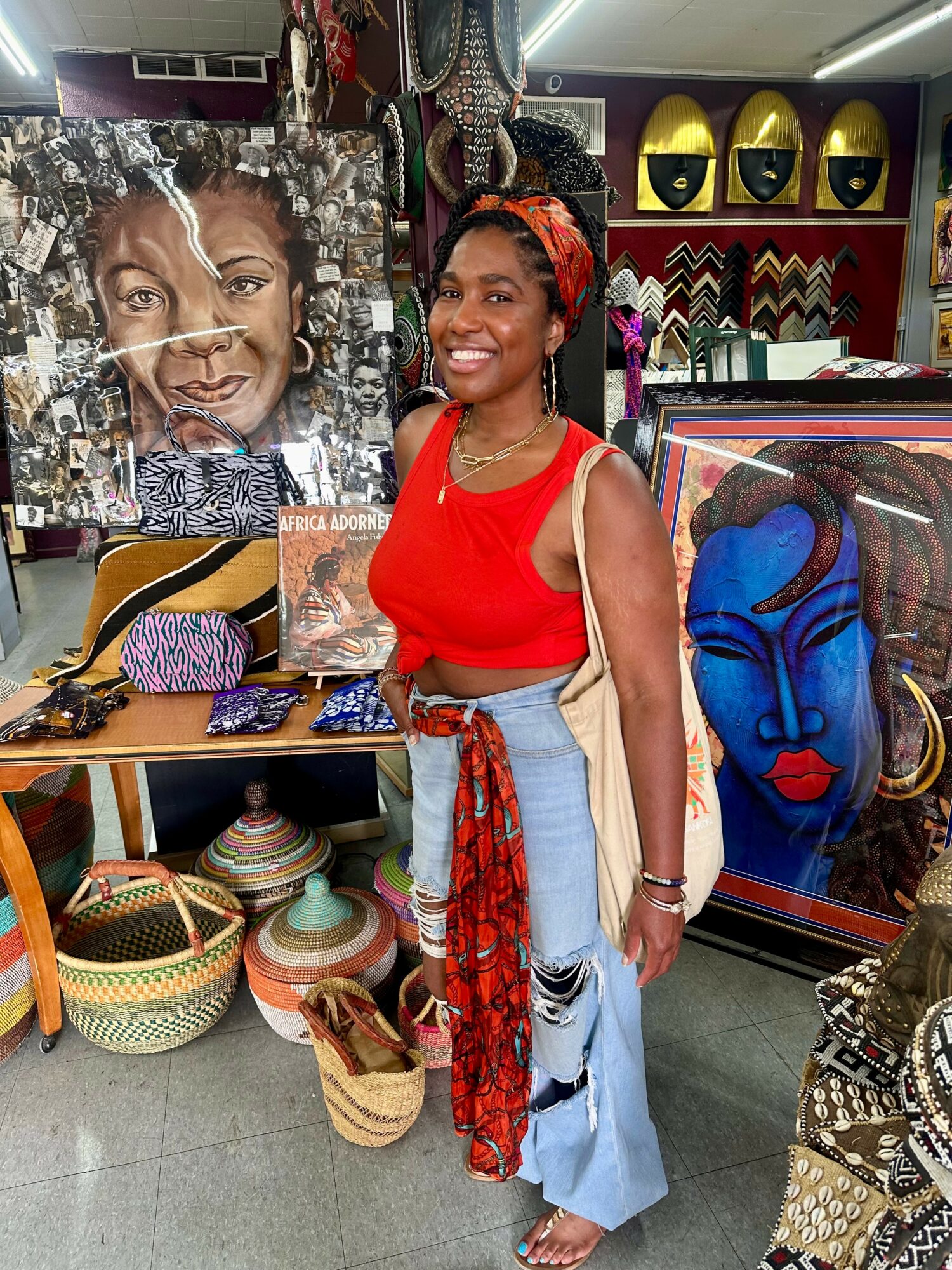 Smiling woman in red top and light blue jeans stands in art and craft store with paintings and woven baskets.