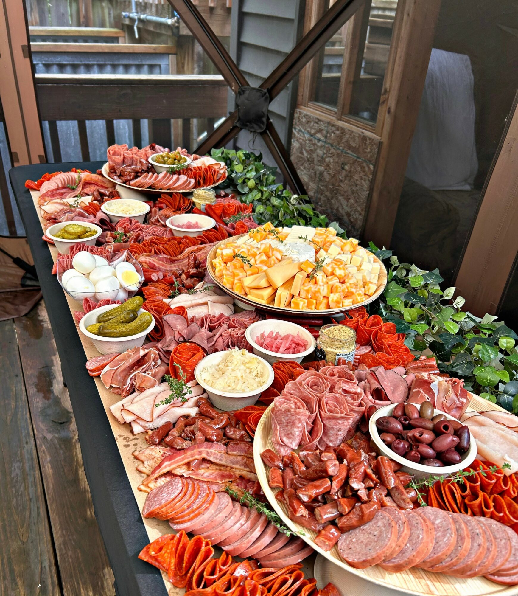 A table with various meats, cheeses, olives, and vegetables arranged for a meal outdoors, with a wooden deck and railing in background.