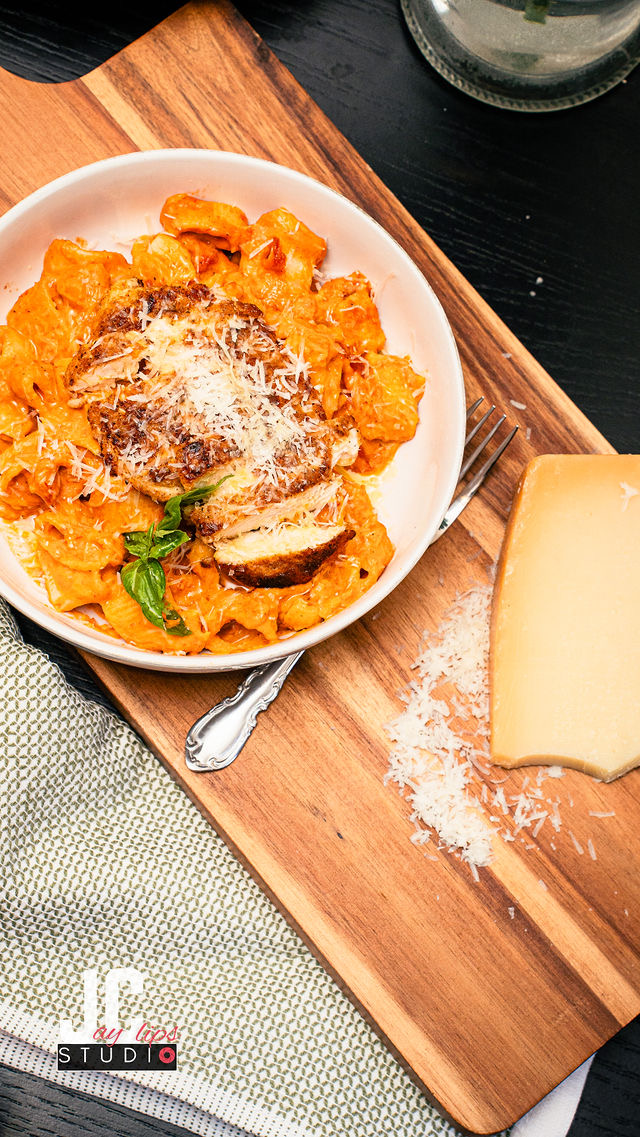 Bowl of pasta with cheese, basil, and vegetables on a wooden board, with cheese and flour nearby, on a table.