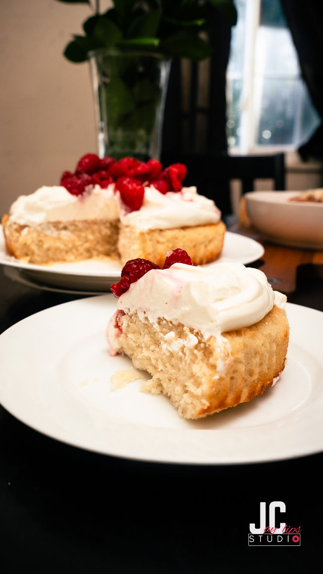 Slice of cake with whipped cream and red berries on a white plate, with a second cake slice in the background.