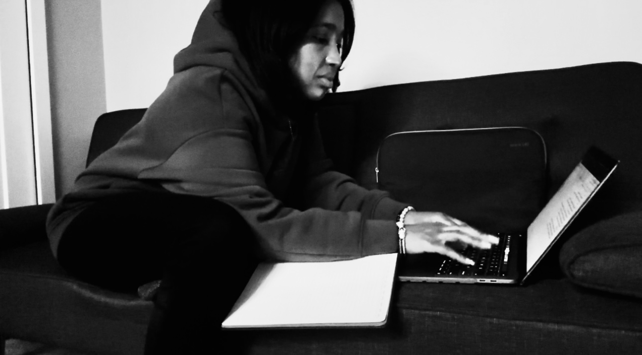 Person sitting on a couch using a laptop, with a closed notebook nearby, in black and white.
