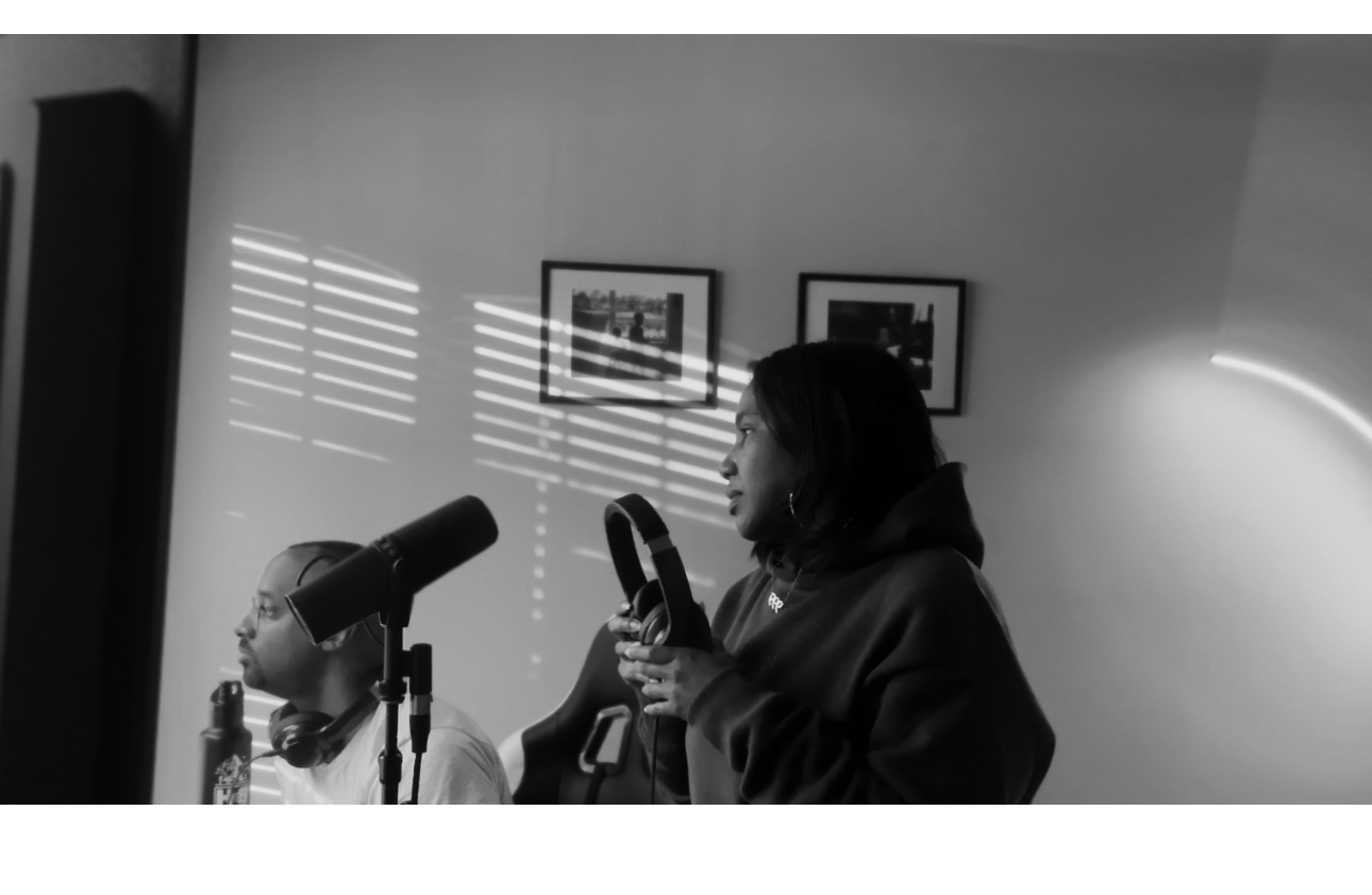 Two women speaking into microphones, one holding a steering wheel, with framed pictures on the wall behind them.