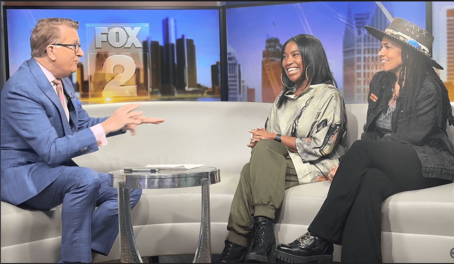 Two women and a man sit on a TV set, engaged in conversation, with a cityscape background and a FOX 2 logo.