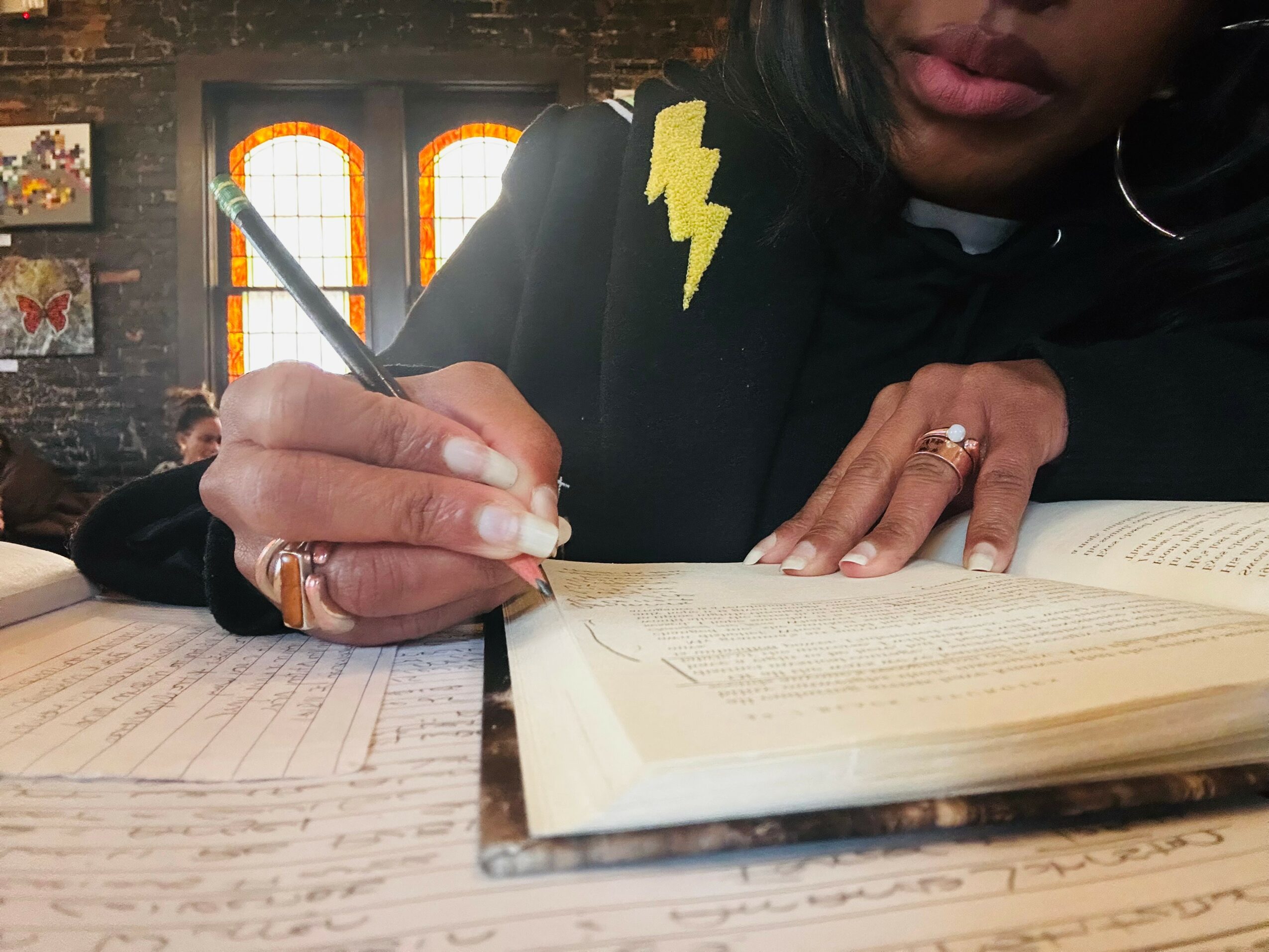 Person writing in a book with a pen, wearing a black top with a yellow lightning bolt patch, in a room with stained glass windows.