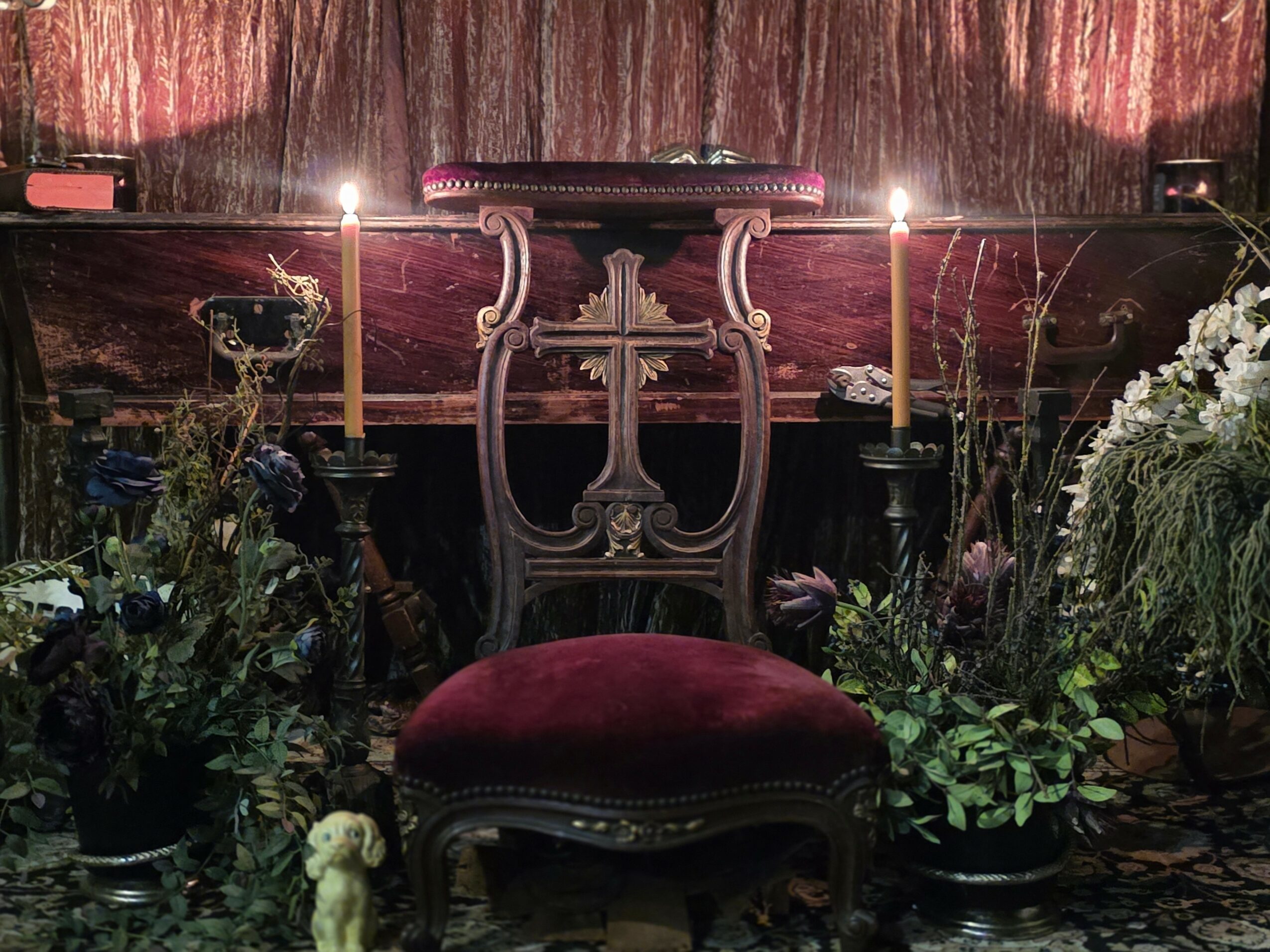 Ornate chair with red velvet cushion surrounded by candles and floral arrangements, set against a dark curtain backdrop.