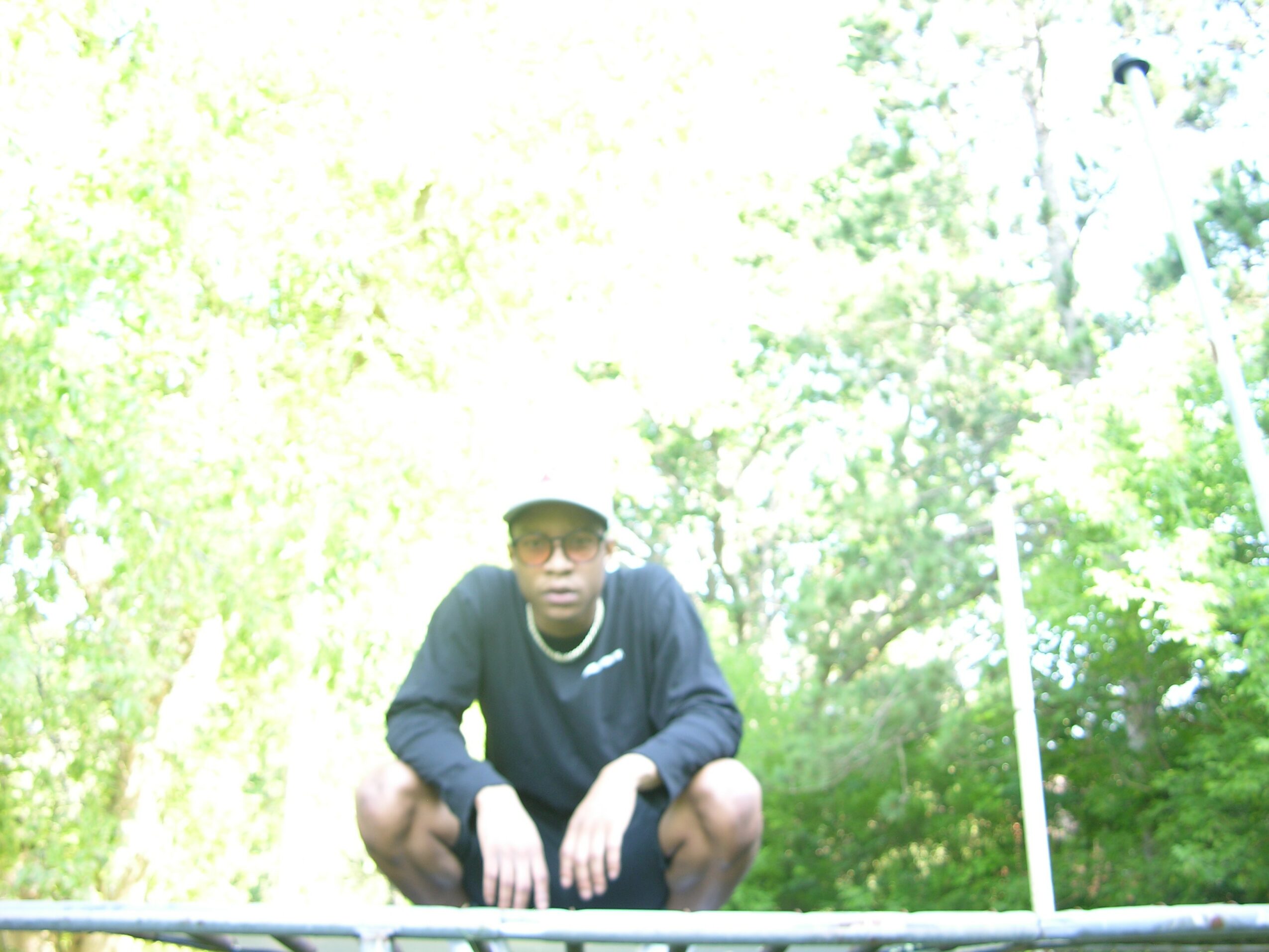 Person crouching outdoors in front of green trees, wearing a dark jacket, cap, and glasses.
