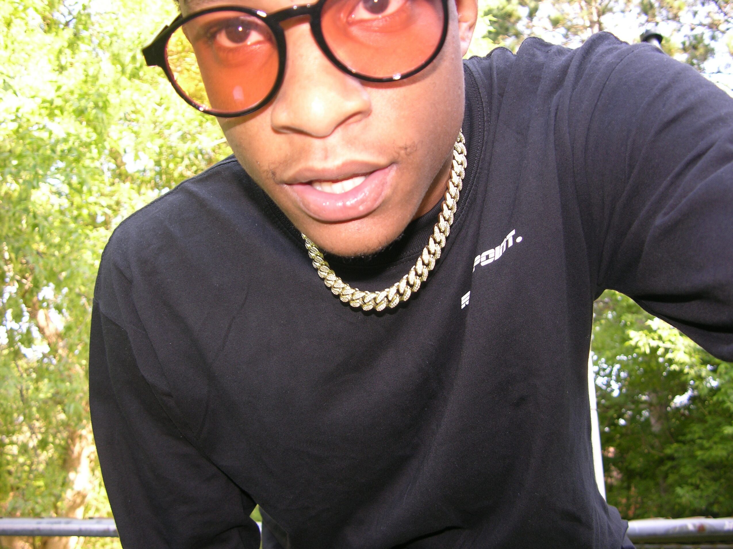Close-up of a person wearing glasses, a chain necklace, and a black shirt outdoors with trees in the background.