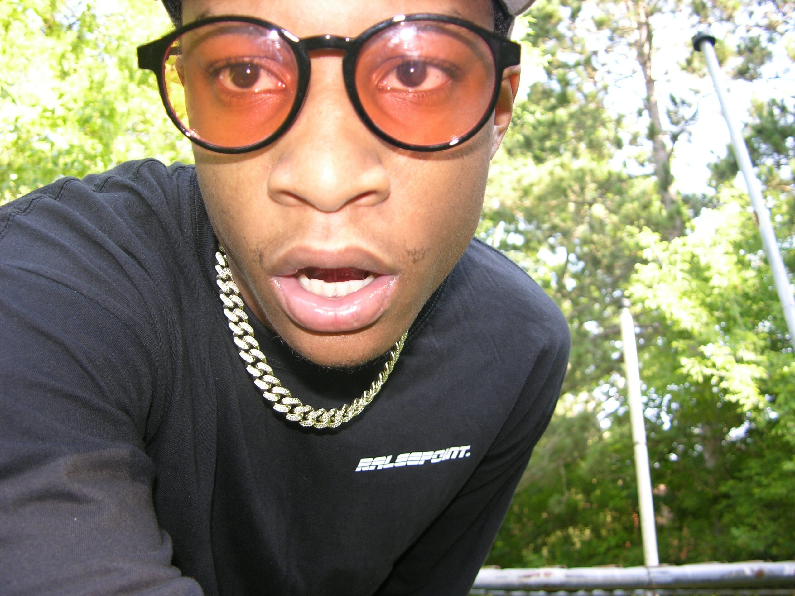 Close-up of a person wearing glasses and a chain, outdoors with trees in the background.