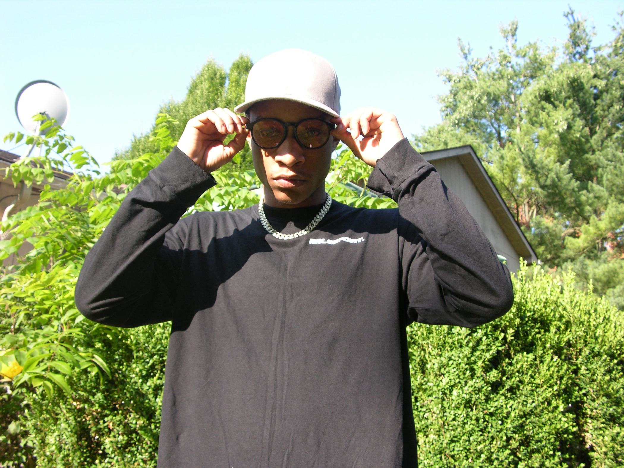 Person wearing glasses, a white hat, and a black long-sleeve shirt outdoors with greenery in the background.